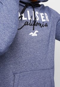 Mörkblå hoodie tillverkad av ett texturerat tyg. Har en kenguruficka, vit tryckt text "Hollister California" och svart dragsko.