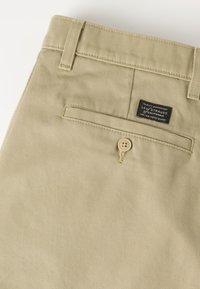 Μπεζ παντελόνι Levi Strauss που δείχνει την πίσω τσέπη με κλείσιμο με κουμπί και μια μαύρη ετικέτα της μάρκας Levi Strauss πάνω από την τσέπη.