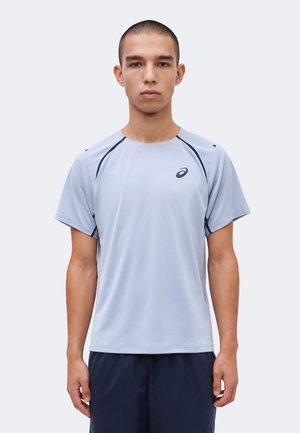 Νέος άνδρας με κοντά μαλλιά, φορώντας ένα αθλητικό ανοιχτό γκρι T-shirt Asics και σκούρα σορτς, στέκεται μπροστά σε απλό φωτεινό φόντο.