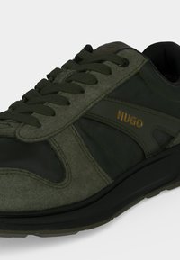 Baskets vert foncé avec des accents noirs et des lacets, portant le nom de la marque Hugo en lettres dorées sur le côté.