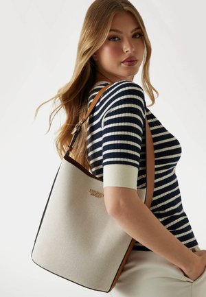 Canvas-Shopper in Weiß mit braunen Lederakzenten und einem Logo. Die Tasche hat eine strukturierte Form und einen langen Schulterriemen.