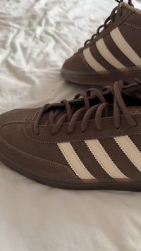 Rjavi semiš športni copati s kremno belimi črtami, okroglega prstnega dela, vezalkami, teksturiranim zgornjim delom in gumijastim podplatom. Prikaz v bližnjem posnetku poudarja podrobnosti.