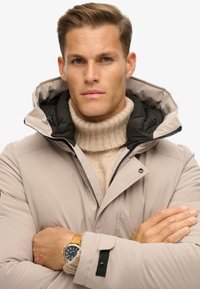 Beige isoleret jakke med hætte, sort indvendig foring og knapdetaljer. Laget med en tan strikket turtleneck. Guld ur synligt.