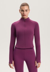 Přiléhavá fialová sportovní bunda s vysokým límcem a plným zipem, dlouhými rukávy a otvory na palce; vyrobená z pružné, hladké tkaniny.