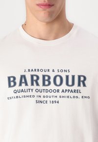 Λευκό κοντομάνικο μπλουζάκι με σκούρο μπλε κείμενο που αναγράφει "BARBOUR" και πρόσθετες λεπτομέρειες εταιρικής ταυτότητας μπροστά. Από βαμβακερό υλικό, σχέδιο με στρογγυλή λαιμόκοψη.