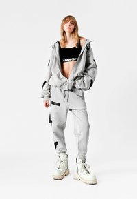 Joven mujer de pie con chaqueta gris claro de cremallera extragrande y pantalones a juego con detalles en negro, top corto negro y botas blancas con cordones.