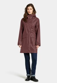 Mauve waterdichte regenjas met capuchon, sluiting met drukknoop aan de voorkant en twee grote zakken aan de voorkant. Gemaakt over donkere jeans en zwarte laarzen.