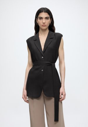 Γυναίκα με μακριά σκοτεινά μαλλιά φοράει αμάνικο μαύρο σακάκι με ζώνη και μπεζ παντελόνια με φαρδιά μπατζάκια μπροστά σε απλό λευκό φόντο.