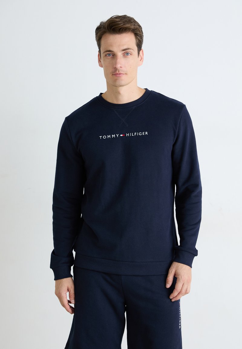 Mies, joka on pukeutunut laivastonsiniseen Tommy Hilfiger -huppariin ja samanvärisiin shortseihin, seisoo yksinkertaista valkoista taustaa vasten.