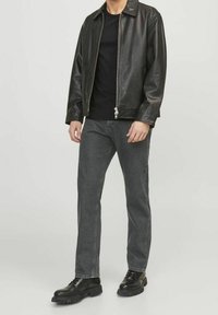 Chaqueta de cuero negra con cuello, cierre de cremallera y mangas largas. Usada sobre una camisa negra, combinada con jeans gris oscuro y botas negras.