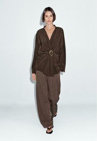 Blouse en satin marron avec un détail de nœud à l'avant et un accent en anneau doré, associée à un pantalon large marron. Modèle portant des sandales noires.