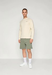 Lys beige sweatshirt med ribbestrikkede mansjetter, kombinert med olivengrønne shorts. Modellen har på seg hvite joggesko og ankelsokker. Enkel design.