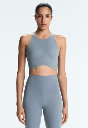 Kvinde iført en matchende lyseblå tætsiddende crop top og højttaljede leggings, stående med armene afslappet ned langs kroppen mod en ensfarvet baggrund.