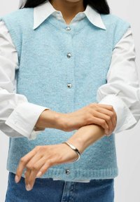 Lyseblå strikket vest med v-hals, sølvfargede knappelukkinger og teksturert overflate, lagdelt over en hvit skjorte med oppbrettede mansjetter.