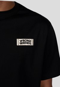 Czarna bawełniana koszulka z krótkim rękawem, z okrągłym dekoltem, z prostokątną, białą metką z czarnym napisem "WRONG FRIENDS". Gładka tekstura.