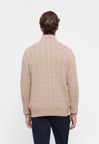 Man met de rug naar de camera, gekleed in een beige kabelgebreide trui met een geribbelde kraag, manchetten en onderrand, in combinatie met donkerblauwe broek tegen een witte achtergrond.