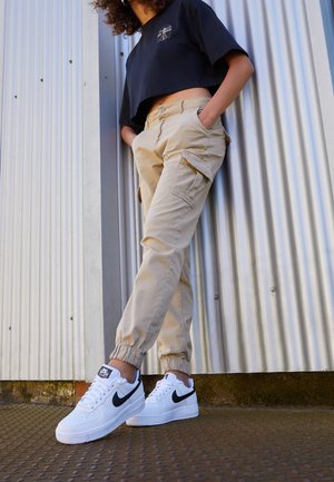 Personne portant un pantalon cargo beige, un t-shirt noir court et des baskets blanches Nike Air Force 1, debout contre un mur en métal ondulé.