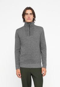 Maglione grigio con zip, realizzato in un design a maglia testurizzata, colletto alto e orlo a coste. Indossato con pantaloni verde scuro.