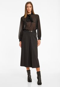 Schwarze durchsichtige Bluse mit einem Schalkragen und Schleifenverschluss, kombiniert mit hoch taillierten schwarz-weiß gestreiften Culottes und schwarzen Knöchelstiefeln.