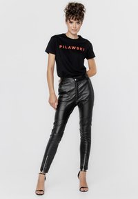 Czarna skrócona koszulka z napisem "PILAWSKI" w czerwonym kolorze. Czarne spodnie z wysokim stanem z syntetycznej skóry z zamkami na mankietach i gładką fakturą. Sandały na wysokim obcasie.