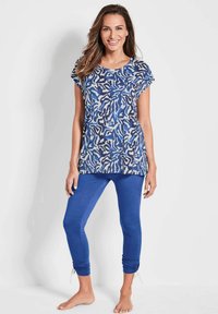 Vrouw die glimlacht, gekleed in een blauw gestructureerd t-shirt met korte mouwen en effen blauwe gerimpelde leggings, staande op blote voeten tegen een witte achtergrond.