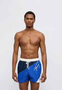 Μπλε και ναυτικό σορτς για κολύμβηση με ελαστική μέση, κορδόνι κ αι λεπτομέρεια με λογότυπο. Κατασκευασμένα από ελαφρύ ύφασμα, με σχέδιο σε μπλοκ χρωμάτων.