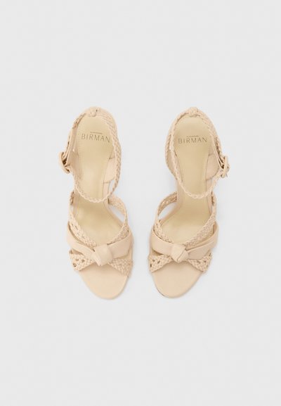 Μπεζ σανδάλια με ψηλό τακούνι και λουράκια, με πλεκτές λεπτομέρειες και λουράκια στον αστράγαλο, τοποθετημένα επίπεδα σε ανοιχτό φόντο.