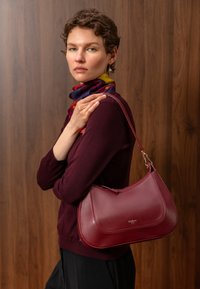 Femme aux cheveux courts portant un pull bordeaux et une écharpe colorée, tenant un sac à main en cuir rouge sur son épaule devant un fond en bois.