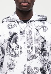 Άτομο με σκουρόχρωμο δέρμα που φοράει λευκό φούτερ με φερμουάρ, διακοσμημένο με μαύρα και γκρι στροβιλιζόμενα μπαρόκ και πιτσιλιές μοτίβα.