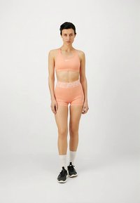 Brassière de sport pêche claire et short taille haute avec logo Nike blanc, assortis à des chaussures de sport noires et des chaussettes blanches. Texture de tissu lisse.