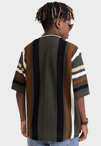 Knit polo shirt with green, brown, white, and black stripes. Short sleeves, ribbed collar, and loose fit. Textured knit pattern visible.