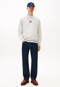 Γκρι φούτερ με κόκκινο και μπλε λογότυπο, σκούρο μπλε τζιν παντελόνι και καφέ δερμάτινα παπούτσια με κορδόνια, συνδυασμένα με καπέλο σε navy μπλε απόχρωση.