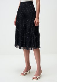Gonna midi nera plissettata con motivo ricamato a stelle, vita elastica e sovrapposizione trasparente, abbinata a sandali con tacco aperto bianchi.