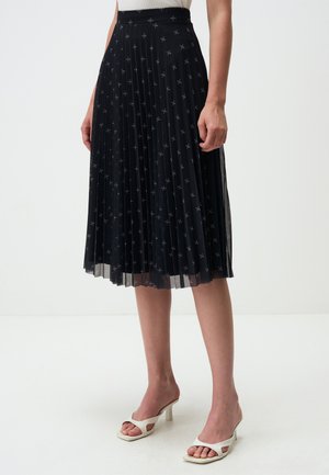 Jupe midi plissée noire avec motif étoile brodé, taille élastique et voilage transparent, associée à des sandales à talons ouverts blanches.