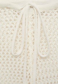 Gilet en tricot crème, avec un motif en maille lâche, noué à la taille avec des liens en tissu plats et assortis. Texture douce et design léger.