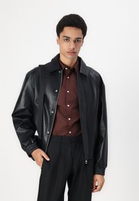 Giacca nera in ecopelle con zip frontale, polsini elasticizzati e colletto appuntito, indossata sopra una camicia marrone e pantaloni neri.