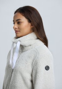 Nainen, jolla on pitkät ruskeat hiukset, pukeutuneena valkoiseen sherpa-fleece-takkiin, jossa on korkea kaulus ja nyörit, kasvoillaan vasemmalle yksinkertaista taustaa vasten.