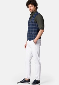 Granatowa kamizelka w kratę z pięcioma guzikami na ciemnozielonej koszuli, zestawiona z białymi spodniami i granatowymi sneakersami z białymi podeszwami.