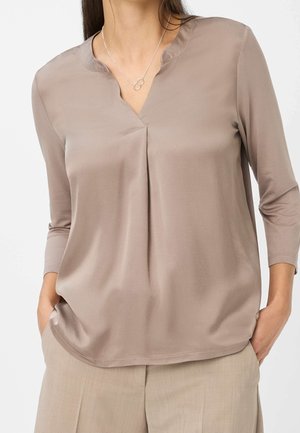 Kobieta w taupe satynowej bluzce z rękawami do połowy przedramienia i srebrnym naszyjnikiem z przeplatającymi się kółkami, z rękami w kieszeniach beżowych spodni.