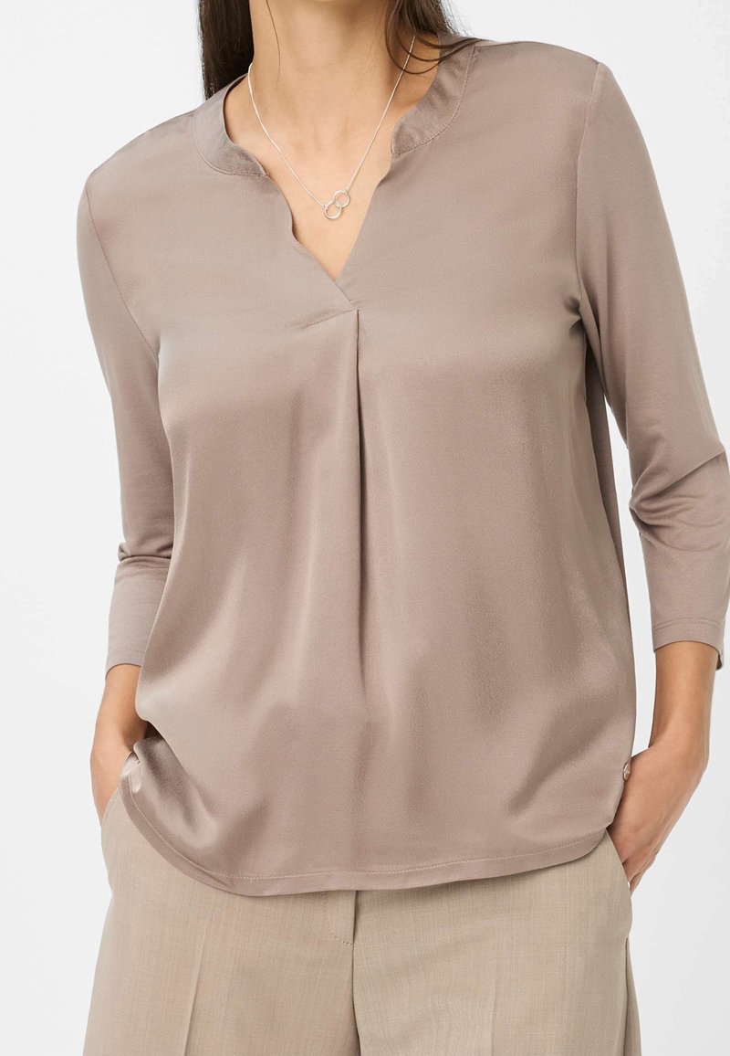 Femme portant un chemisier en satin taupe à manches trois-quarts et un collier argenté avec des cercles entrelacés, les mains dans les poches d'un pantalon beige.