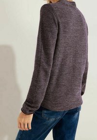 Langärmliges Shirt aus dunkel meliertem lila Stoff mit leichtem Muster, Rundhalsausschnitt und taillierter Passform. Kombiniert mit blauen Jeans.