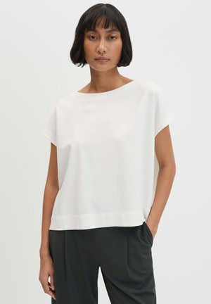 Frau mit kurzen schwarzen Haaren, die ein lockeres weißes kurzärmeliges Oberteil und dunkle Hosen trägt, steht mit einer Hand in der Tasche.