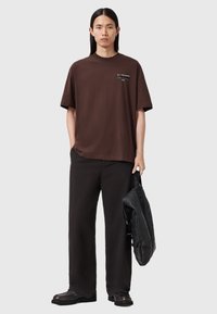 Braunes Baumwoll-T-Shirt mit lockerem Schnitt, kurzen Ärmeln und einem weißen Logo auf der Brust. Kombiniert mit schwarzen, weit geschnittenen Hosen und einer schwarzen Lederhandtasche.