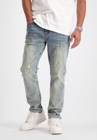 Lichtblauwe versleten jeans met scheuren en vervaging, gedragen met een wit T-shirt en witte sneakers. De denim heeft een relaxte pasvorm en taps toelopende pijpen.