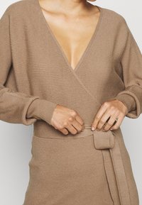 Robe portefeuille beige en tricot avec un col en V profond, une texture côtelée et des manches longues. Comprend une ceinture nouée en tissu assorti à la taille.