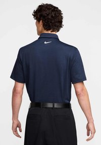 Jūrinio stiliaus polo marškiniai su trumpomis rankovėmis ir apykakle, su balta Nike logotipu nugaroje, derinami su juodomis kelnėmis ir juodu diržu.