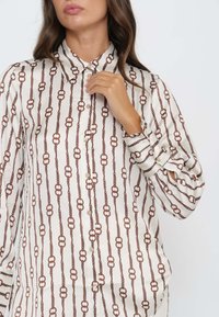 Hemd mit Knopfleiste aus glattem Stoff, das ein Kettenmuster in Braun auf einem hellbeigen Hintergrund aufweist. Enthält einen schmalen Kragen und geschlossene Ärmel.