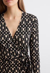 Robe noire portefeuille à manches longues, ornée d'un imprimé géométrique beige, décolleté en V et détail de taille nouée. Tissu lisse et léger.