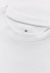 Artimas baltos medvilnės marškinėlių su apykakle vaizdas, kuriame matyti vidutinio dydžio etiketas apykaklėje.