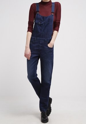 Henkilö, joka pukeutunut tummansinisiin farkkuhaalaritaskuihin ja marjapuuronväriseen ribbineuletoppiin, seisoo yhdellä kädellä taskussa ja mustissa kengissä.